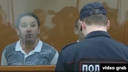 Яке аз маҳкумшудаҳо аз пушти оинаи курсии айбдоршавандаҳо истода аз худ дифоъ мекард. Акс аз навори видеоии NTV гирифта шудааст 